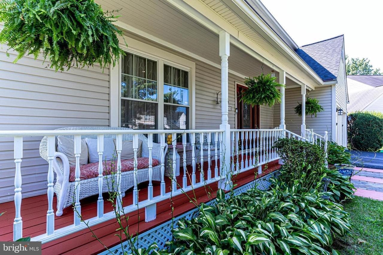 The view of a home with a long front porch with red planks, white railing, white wicker furniture, and plants out front
